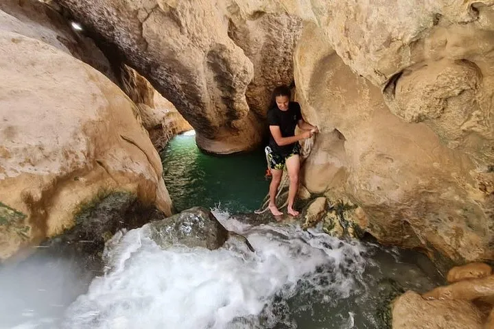 A swimmer climbing through a narrow gap in the rock beside a small cascade in Wadi Shab