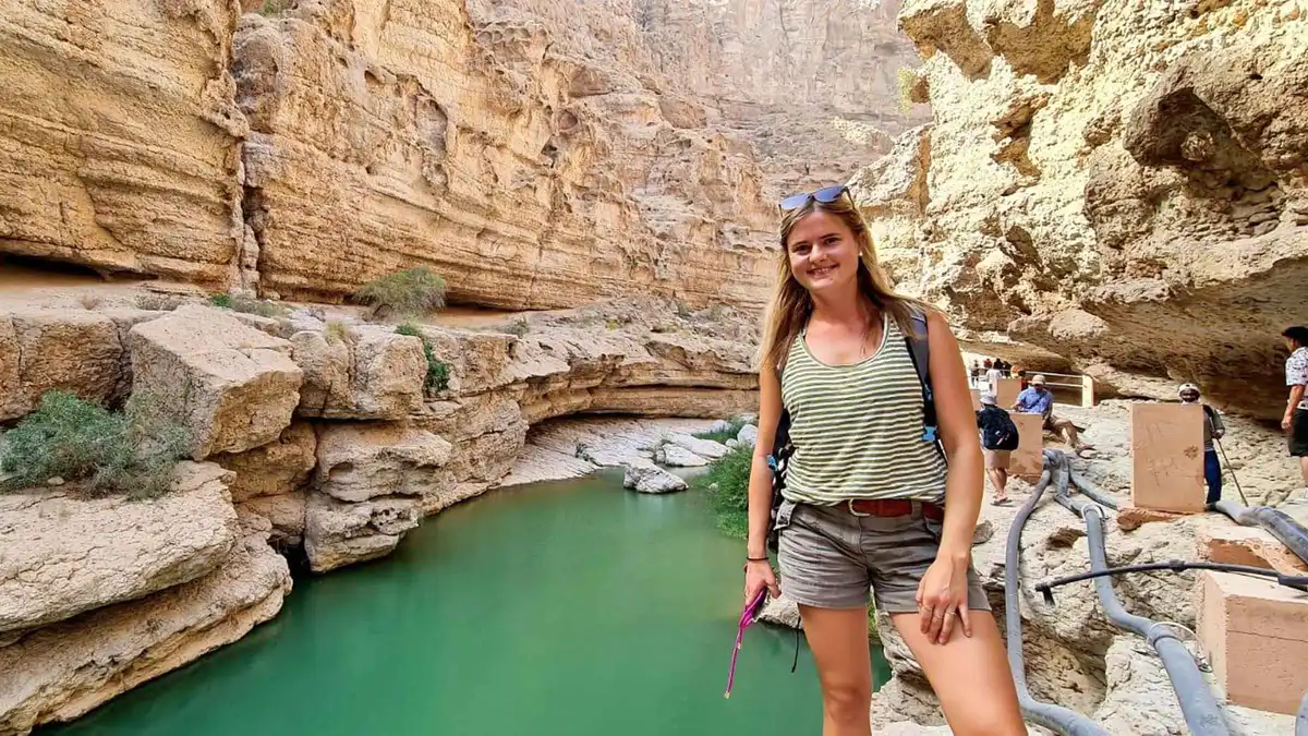 A hiker on the path beside a green pool and stepped rock ledges in Wadi Shab